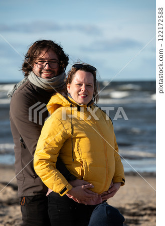 A couple stands embraced on a sandy beach on a chilly autumn day, the serene ocean stretching behind them as they share a quiet moment of love and warmth amidst the cool breeze and peaceful 122172588