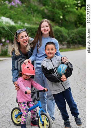 A happy group of people, including two women, a boy holding a ball, and a little girl on a balance bike with a helmet, enjoying outdoor activities on a paved path surrounded by greenery. 122172703