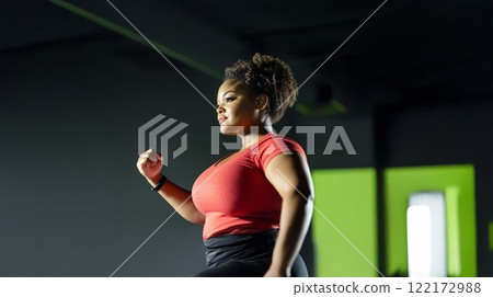 Plus size black woman in sportswear pushing through a workout at the gym, sweat dripping from her focused face. Generated image 122172988