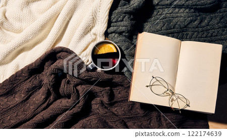 Aerial view of open book and glasses resting on knitted blankets with hot drink in metal mug with orange slice, representing winter relaxation and creative inspiration. Aerial view of open book and glasses resting on knitted blankets with hot drink in metal mug with orange slice, representing winter relaxation and creative inspiration. 122174639