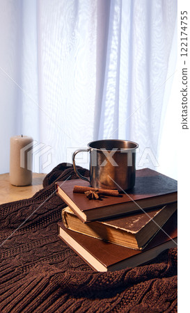 Metal mug on stack of vintage books with cinnamon sticks on soft brown knitted blanket, candle glowing softly in background for winter ambiance. 122174755