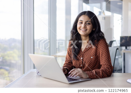 Portrait young successful woman at her workplace inside the office. Businesswoman smiling and looking at camera, typing on the laptop keyboard, sitting by the window, happy and satisfied employee Portrait young successful woman at her workplace inside the office. Businesswoman smiling and looking at camera, typing on the laptop keyboard, sitting by the window, happy and satisfied employee 122174965