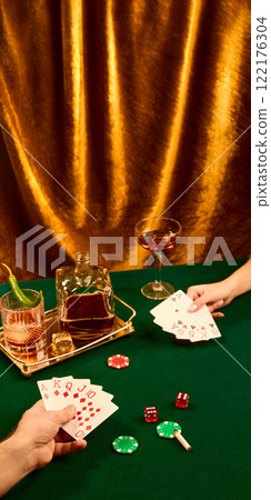 Still life photography, Man and woman playing cards, sitting at table with whiskey glasses, poker chips and dice against golden curtain. Luxury and drama 122176304