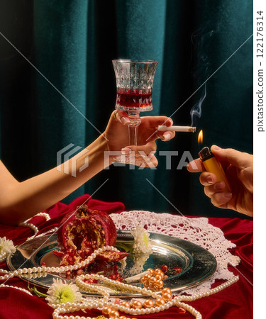 Female hand holding wine glass and cigarette, surrounded by split pomegranate, pearls, and white flowers on silver tray against green velvet background. Female hand holding wine glass and cigarette, surrounded by split pomegranate, pearls, and white flowers on silver tray against green velvet background. 122176314