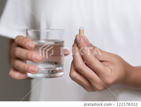 Woman in white t-shirt holding beige capsule in fingers and water glass in hand closeup Woman in white t-shirt holding beige capsule in fingers and water glass in hand closeup 122176332