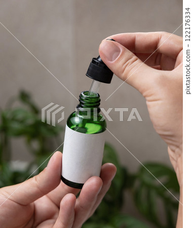 Closeup of female hands opening green dropper bottle with blank label, cosmetic mockup 122176334