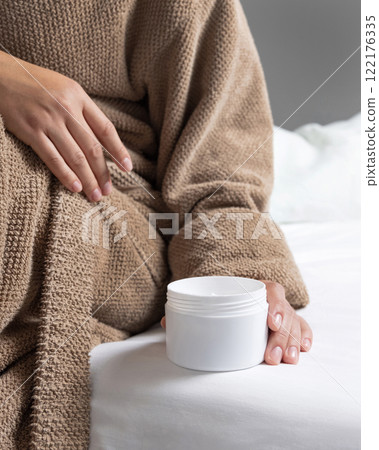 Woman in brown bathrobe puts white opened cream jar on white bedsheet closeup, mockup Woman in brown bathrobe puts white opened cream jar on white bedsheet closeup, mockup 122176335