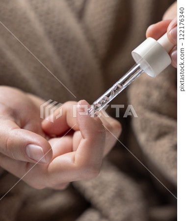 Woman in brown bathrobe dropping cuticle oil on a fingernail and holding a pipette closeup Woman in brown bathrobe dropping cuticle oil on a fingernail and holding a pipette closeup 122176340
