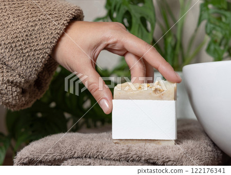 Closeup of female hand in brown bathrobe holding soap bar with blank label, mockup Closeup of female hand in brown bathrobe holding soap bar with blank label, mockup 122176341