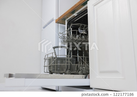 Side view of an empty open dishwasher with gray drawers for dishes and cutlery stands against the background of white kitchen cabinets and a white refrigerator. Dishwashing concept, comfort. High Side view of an empty open dishwasher with gray drawers for dishes and cutlery stands against the background of white kitchen cabinets and a white refrigerator. Dishwashing concept, comfort. High 122177186