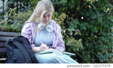 A teenage girl studies outdoors, pen in hand and books open, with a backpack beside her. This photo captures a peaceful moment of learning in a natural setting. 122177279