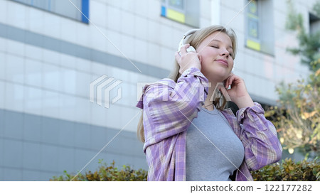 A young woman listens to music on her headphones in an outdoor urban setting, smiling softly. This photo showcases casual confidence and the joy of everyday moments. A young woman listens to music on her headphones in an outdoor urban setting, smiling softly. This photo showcases casual confidence and the joy of everyday moments. 122177282