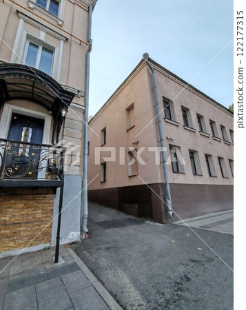 Two contrasting buildings meet at a narrow alleyway: a decorative beige facade with ornate ironwork and a plain light pink structure. The photo highlights architectural contrast and urban geometry Two contrasting buildings meet at a narrow alleyway: a decorative beige facade with ornate ironwork and a plain light pink structure. The photo highlights architectural contrast and urban geometry 122177315