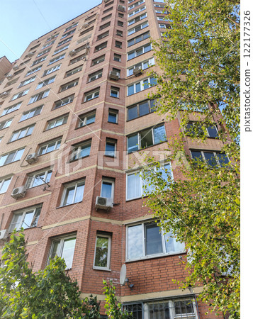 A tall red and beige brick residential building with multiple balconies, air conditioning units, and a sidewalk below. The structure reflects the typical architecture of urban apartment blocks in 122177326