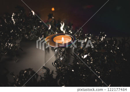 Burning candle in metal candle holder amongst silver tinsel for Christmas tree on dark background. Fire, candle flame. Festive party decor. Burning candle in metal candle holder amongst silver tinsel for Christmas tree on dark background. Fire, candle flame. Festive party decor. 122177484