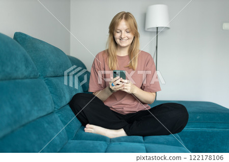 Beautiful blonde woman having fun online on her smartphone at home or hotel. Phone Communication and comfort concept. Smiling 30s woman daydreaming and holding mobile phone with copy space. Full body Beautiful blonde woman having fun online on her smartphone at home or hotel. Phone Communication and comfort concept. Smiling 30s woman daydreaming and holding mobile phone with copy space. Full body 122178106