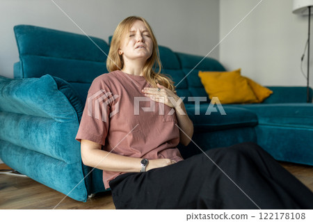 Woman sitting with strong chest pain and hand touching her chest while having trouble at home. Heart attack concept or heart failure symptom. Pressure in the chest. 122178108