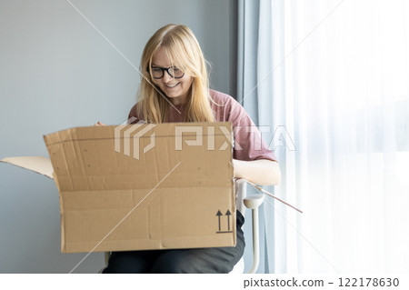 Young cheerful woman in glasses getting parcel at home. Pretty girl sitting on sofa at home and opening carton box. Express delivery service and satisfied consumer. Online shopping banner Young cheerful woman in glasses getting parcel at home. Pretty girl sitting on sofa at home and opening carton box. Express delivery service and satisfied consumer. Online shopping banner 122178630