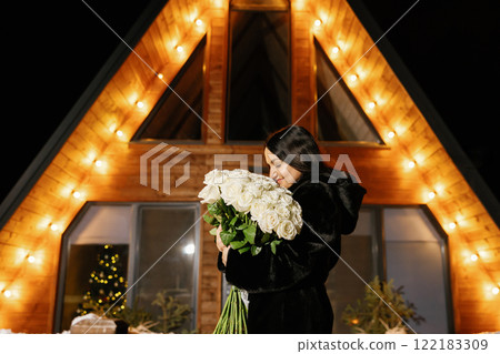 Woman holding white roses bouquet in front of illuminated wooden house 122183309