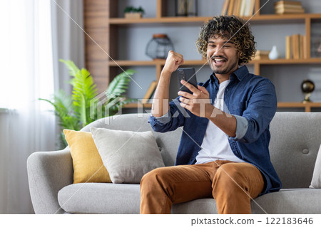 Successful joyful man sitting at home on sofa with phone in hands, celebrating victory and successful achievement results. Freelancer received online notification with good news on smartphone. 122183646