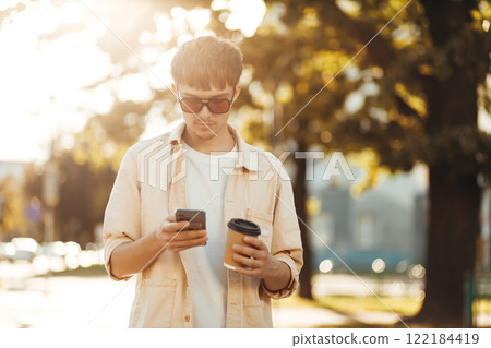 Smiling Young Man Texting on Smartphone and Holding Coffee in Morning City Street Smiling Young Man Texting on Smartphone and Holding Coffee in Morning City Street 122184419
