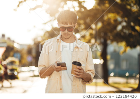 Smiling Young Man Texting on Smartphone and Holding Coffee in Morning City Street 122184420