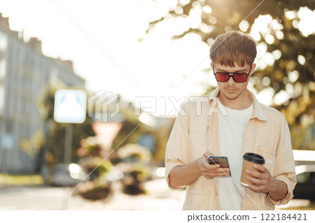 Smiling Young Man Texting on Smartphone and Holding Coffee in Morning City Street 122184421