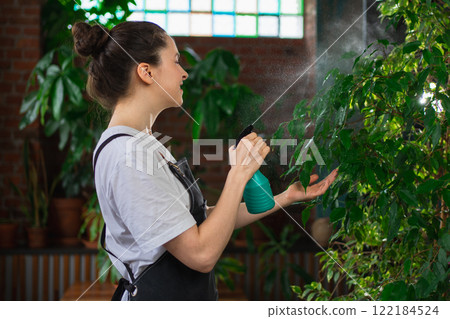 Running of own business. Young woman florist with spray bottle watering fertilizing plants in botanical store. Small business owner working at flower shop smiling surrounded by plants Small business 122184524