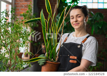 Running of own business. Young woman florist holding plant in pot wearing apron in botanical store. Happy small business owner working at flower shop smiling surrounded by plants. Small business 122184528