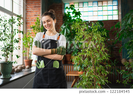 Running of own business. Young woman florist wearing apron in botanical store with green plants. Happy small business owner working at flower shop standing surrounded by plants. Small business 122184545