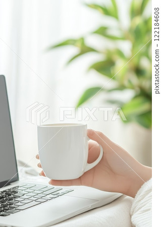 Hands holding a cup of coffee, wrapped in a blanket, with a laptop and oranges in the background, representing comfort, productivity, and warmth 122184608
