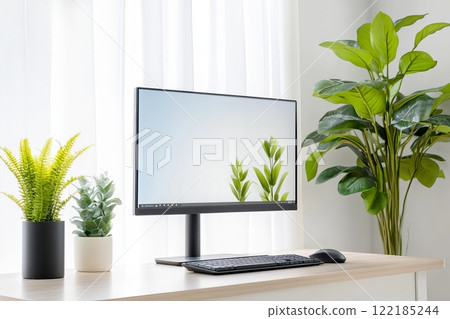 A white desk with a large computer monitor surrounded by indoor plants, books, and stationary, representing a clean and organized workspace 122185244