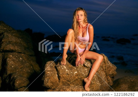Stunning model in bikini sits on ocean beach rocks at night. Sensual woman poses in evening light. Perfect for dating apps or fashion ads. Captures allure, attraction, and feminine charm. Stunning model in bikini sits on ocean beach rocks at night. Sensual woman poses in evening light. Perfect for dating apps or fashion ads. Captures allure, attraction, and feminine charm. 122186292