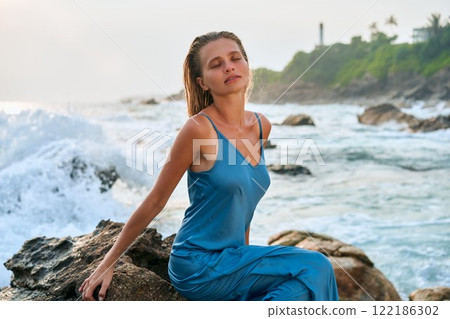 Blonde model in wet dress on cliff with ocean waves and exotic island. Her elegance embodies divine beauty and femininity in nature. Perfect for fashion, travel, nature, or lifestyle themes. Blonde model in wet dress on cliff with ocean waves and exotic island. Her elegance embodies divine beauty and femininity in nature. Perfect for fashion, travel, nature, or lifestyle themes. 122186302