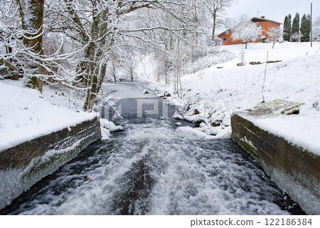Snowy river flowing through a winter landscape with snow-covered banks and trees. 122186384