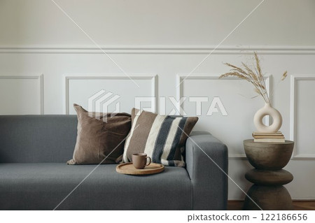 Grey sofa with brown pillows. Empty beige wall mockup. Cup of tea, coffee on wooden tray. Boho living room interior. Modern vase with dry flowers grass. Minimalist home decor. Scandinavian design 122186656