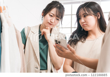 Women and friends choosing clothes while looking at their smartphones at a clothing store/apparel shop (shopping/shopping) Women and friends choosing clothes while looking at their smartphones at a clothing store/apparel shop (shopping/shopping) 122188885