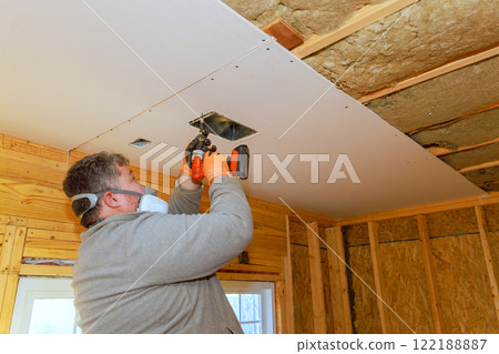 Worker carefully attaches drywall to ceiling while using power screwdriver in an unfinished room. Worker carefully attaches drywall to ceiling while using power screwdriver in an unfinished room. 122188887