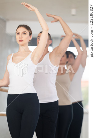 Group of positive young women and men engaged in classical ballet in dance studio performs exercise standing in 3rd position of ballet stance 122190827