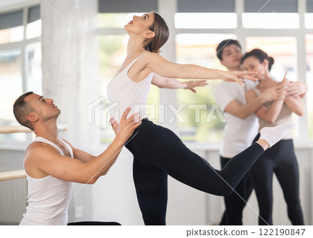 Portrait of positive young man and female partner training ballet pose with support with group of guys and girls in dance class 122190847