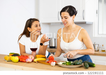 Happy female friends with glasses of red wine chatting and preparing salad together in kitchen Happy female friends with glasses of red wine chatting and preparing salad together in kitchen 122191111