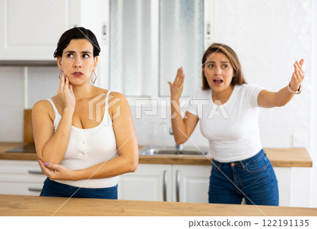 Frustrated young woman listening to angry girlfriend in home kitchen Frustrated young woman listening to angry girlfriend in home kitchen 122191135