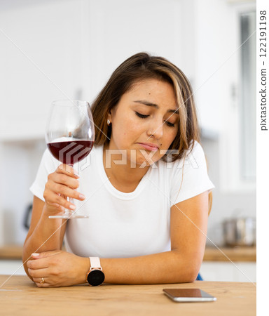Young depressed Latina with glass of wine and silent phone in kitchen 122191198