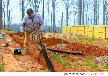 Worker works diligently to install drainage system in his backyard, surrounded by trees fence for enhance water flow management Worker works diligently to install drainage system in his backyard, surrounded by trees fence for enhance water flow management 122191490
