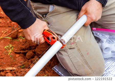 Plumber is using cutting tool to accurately shorten PVC pipe in dirt area during plumbing water line installation 122191522