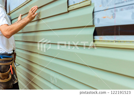 Construction worker carefully aligns, installs green pvc siding board on exterior of house in during renovation project. 122191523