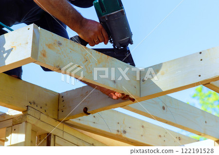 Construction worker is fastening wooden beams using pneumatic nail gun on building site under construction 122191528