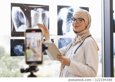 Young Muslim woman doctor records online tutorial using x-rays. She wears glasses and hijab while explaining medical concepts on camera. 122194366
