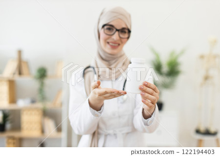 Young Muslim female doctor, in twenties, displaying pill bottle, representing modern medicine and healthcare concept, set in bright clinic environment. 122194403