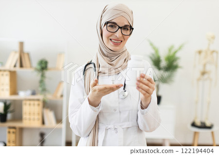 Young Muslim female doctor, in twenties, displaying pill bottle, representing modern medicine and healthcare concept, set in bright clinic environment. 122194404